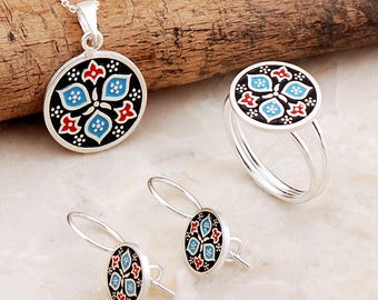 May include: A jewelry set consisting of a circular pendant on a silver chain, a matching ring, and a pair of drop earrings. Each piece features a black enamel background with a symmetrical floral pattern in light blue, red, and white accents.
