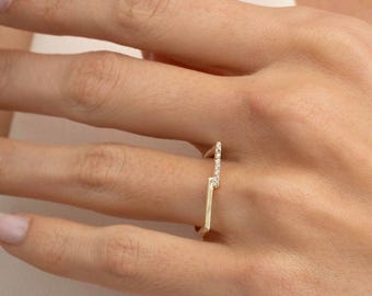 May include: A gold ring featuring a geometric design with two parallel bars. One bar is smooth and plain, while the other is set with a row of small, clear, sparkling stones. The ring has an open, asymmetrical structure that sits across the finger.