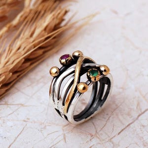 May include: A multi-band ring featuring a combination of silver and gold-toned metal wires that cross over each other. The ring is accented with small gold spheres and two small round gemstones, one red and one green, set into the metal structure.