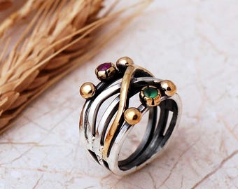 May include: A multi-band ring featuring a combination of silver and gold-toned metal wires that cross over each other. The ring is accented with small gold spheres and two small round gemstones, one red and one green, set into the metal structure.