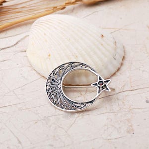 An intricate handmade 925 sterling silver brooch featuring filigree wirework in a crescent moon and star motif, set with a small round red garnet stone and finished with an oxidized antique silver coating.