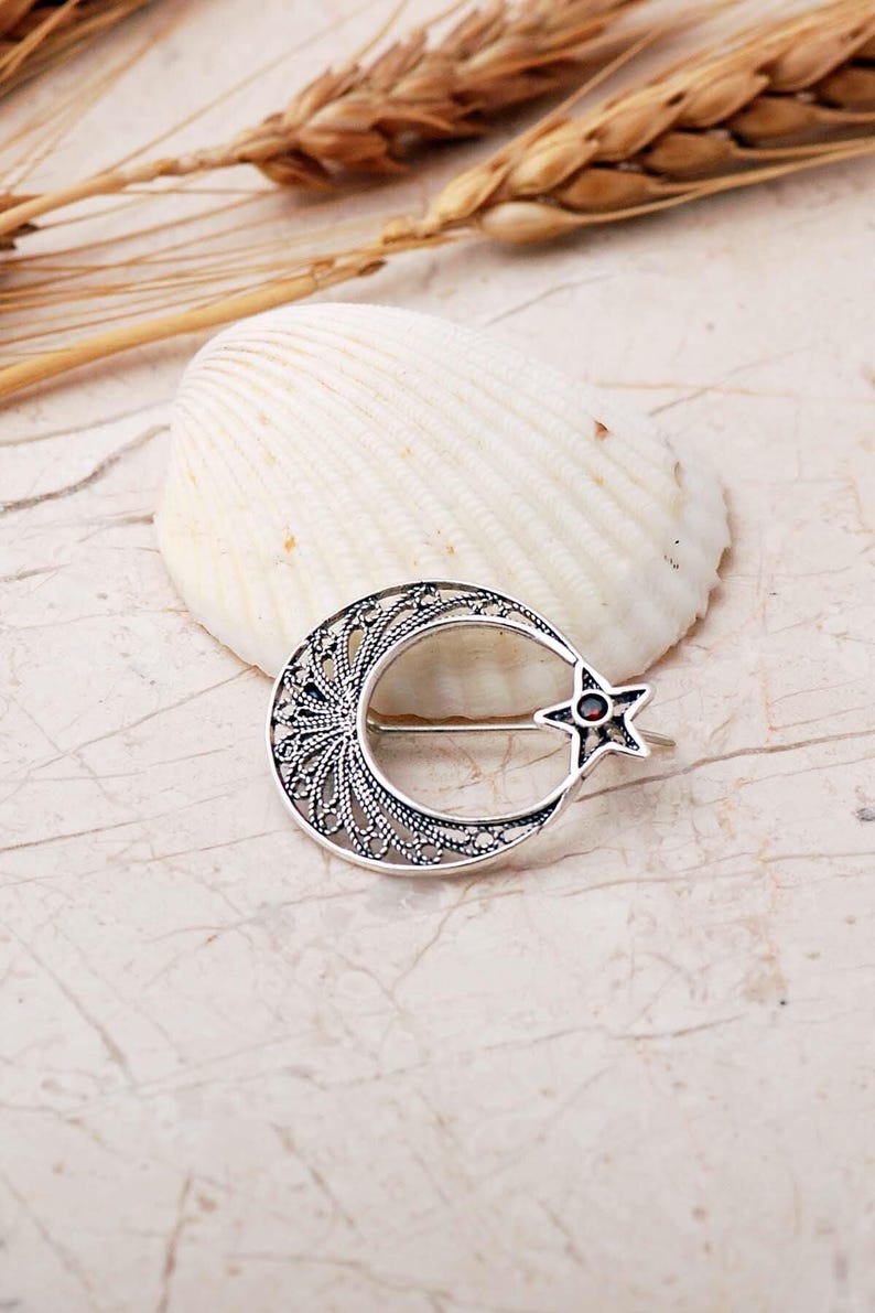 An intricate handmade 925 sterling silver brooch featuring filigree wirework in a crescent moon and star motif, set with a small round red garnet stone and finished with an oxidized antique silver coating.