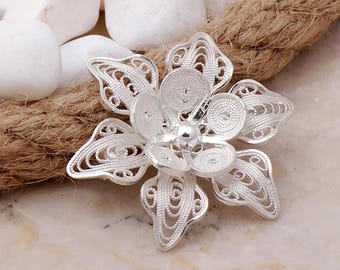 May include: A silver filigree flower brooch with a delicate, intricate design. The flower has a central circle with a raised bead in the centre.
