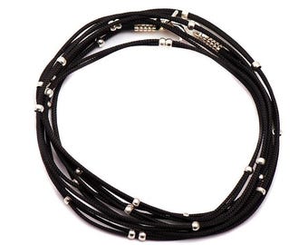 May include: A multi-strand black cord necklace featuring small, polished silver-toned metal beads spaced evenly along the length of the cords. The necklace is arranged in a circular coil and includes a silver-toned metal clasp closure.
