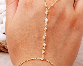 May include: A gold-toned hand chain bracelet featuring a delicate link chain that connects a wrist bracelet to a finger ring. The design is accented with alternating round and pear-shaped clear stones set in gold-toned metal along the center chain.