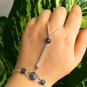 Puede incluir: Una pulsera de mano de plata con un diseño de cadena tejida delicada. La pieza incluye cuatro adornos circulares con centros azul oscuro y patrones florales plateados intrincados, que conectan la pulsera con una presilla para el dedo.