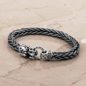 May include: A silver bracelet featuring a braided chain design and a dragon head clasp. The metal has an oxidized finish that highlights the intricate details of the dragon scales and the woven texture of the band, creating a dark, metallic appearance.