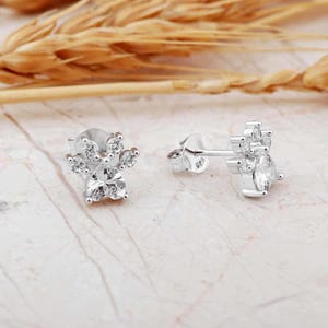May include: A pair of silver stud earrings with a floral design. Each earring features a central, clear, round-cut stone surrounded by smaller, clear stones, creating a sparkling, flower-like appearance. The earrings are set against a light-colored, marble-like surface.