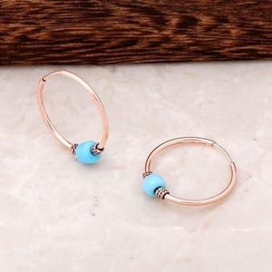 May include: A pair of rose gold hoop earrings, each featuring a single round, bright turquoise bead accented by small, textured metallic spacers on either side of the bead, creating a simple and balanced design for these circular jewelry pieces.