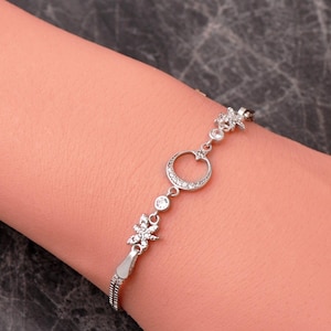 May include: A silver-toned bracelet featuring a central crescent moon charm encrusted with small clear stones, flanked by two star-shaped charms and two round bezel-set stones, all connected by a delicate chain with a sliding adjustable clasp.