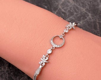 May include: A silver-toned bracelet featuring a central crescent moon charm encrusted with small clear stones, flanked by two star-shaped charms and two round bezel-set stones, all connected by a delicate chain with a sliding adjustable clasp.