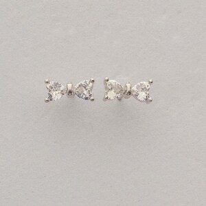 Mini bow‑shaped earrings made of rhodium‑plated sterling silver with a single white zircon stone on each side, measuring 4 mm tall by 1 cm wide.