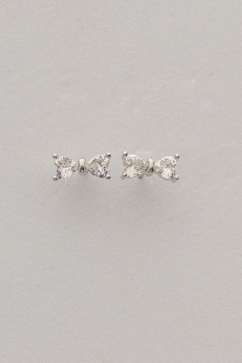 Mini bow‑shaped earrings made of rhodium‑plated sterling silver with a single white zircon stone on each side, measuring 4 mm tall by 1 cm wide.