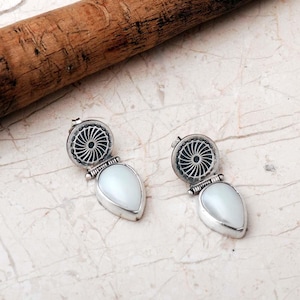 May include: A pair of silver earrings featuring a circular top component with a radial sunburst pattern and a dangling, smooth, white, teardrop-shaped stone. The metal has an oxidized finish in the recessed areas to highlight the intricate details.