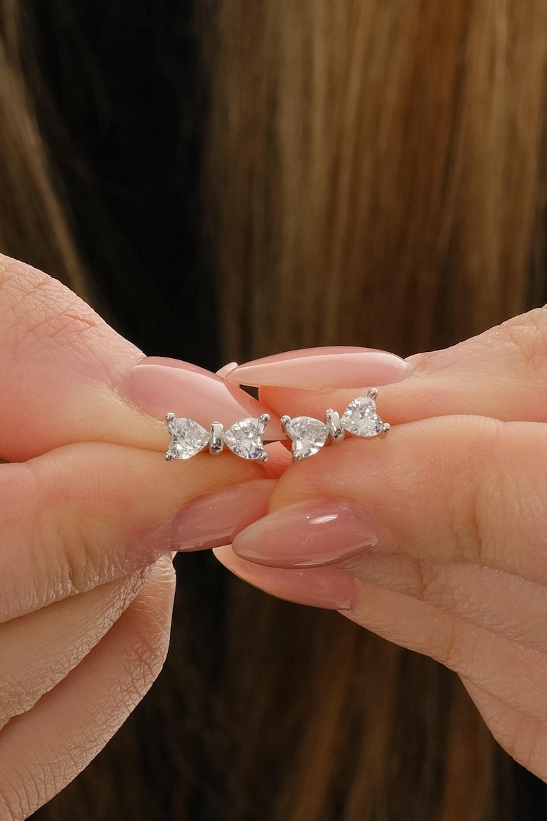 Mini bow‑shaped earrings made of rhodium‑plated sterling silver with a single white zircon stone on each side, measuring 4 mm tall by 1 cm wide.
