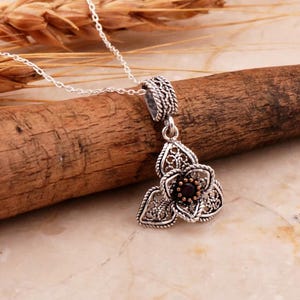May include: A silver necklace with a delicate flower pendant featuring a dark red gemstone in the center. The pendant is made of intricate filigree work.