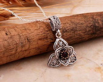 May include: A silver necklace with a delicate flower pendant featuring a dark red gemstone in the center. The pendant is made of intricate filigree work.
