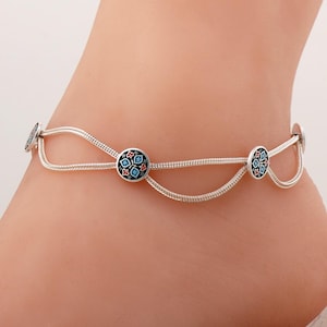 May include: A silver anklet featuring a double-strand chain design with three circular charms. Each charm is decorated with a black enamel background and a floral pattern in red, white, and light blue, creating a delicate and layered appearance.