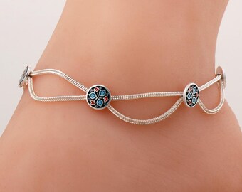 May include: A silver anklet featuring a double-strand chain design with three circular charms. Each charm is decorated with a black enamel background and a floral pattern in red, white, and light blue, creating a delicate and layered appearance.