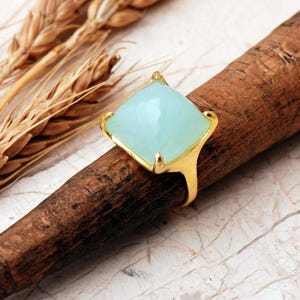 May include: A gold ring featuring a large, square-cut, faceted light blue gemstone set in a four-prong mounting. The stone has a smooth, polished surface with a soft, translucent appearance, and the metal band has a warm, yellow gold finish.