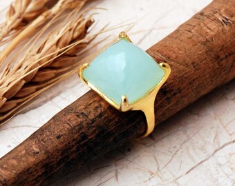 May include: A gold ring featuring a large, square-cut, faceted light blue gemstone set in a four-prong mounting. The stone has a smooth, polished surface with a soft, translucent appearance, and the metal band has a warm, yellow gold finish.