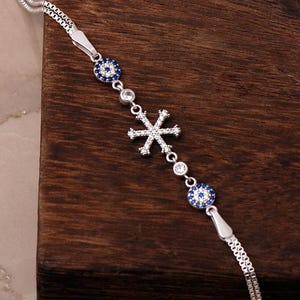 A delicate 925 sterling silver bracelet featuring a sparkling snowflake design combined with a blue sapphire and cubic zirconia evil eye charm on an adjustable box chain.