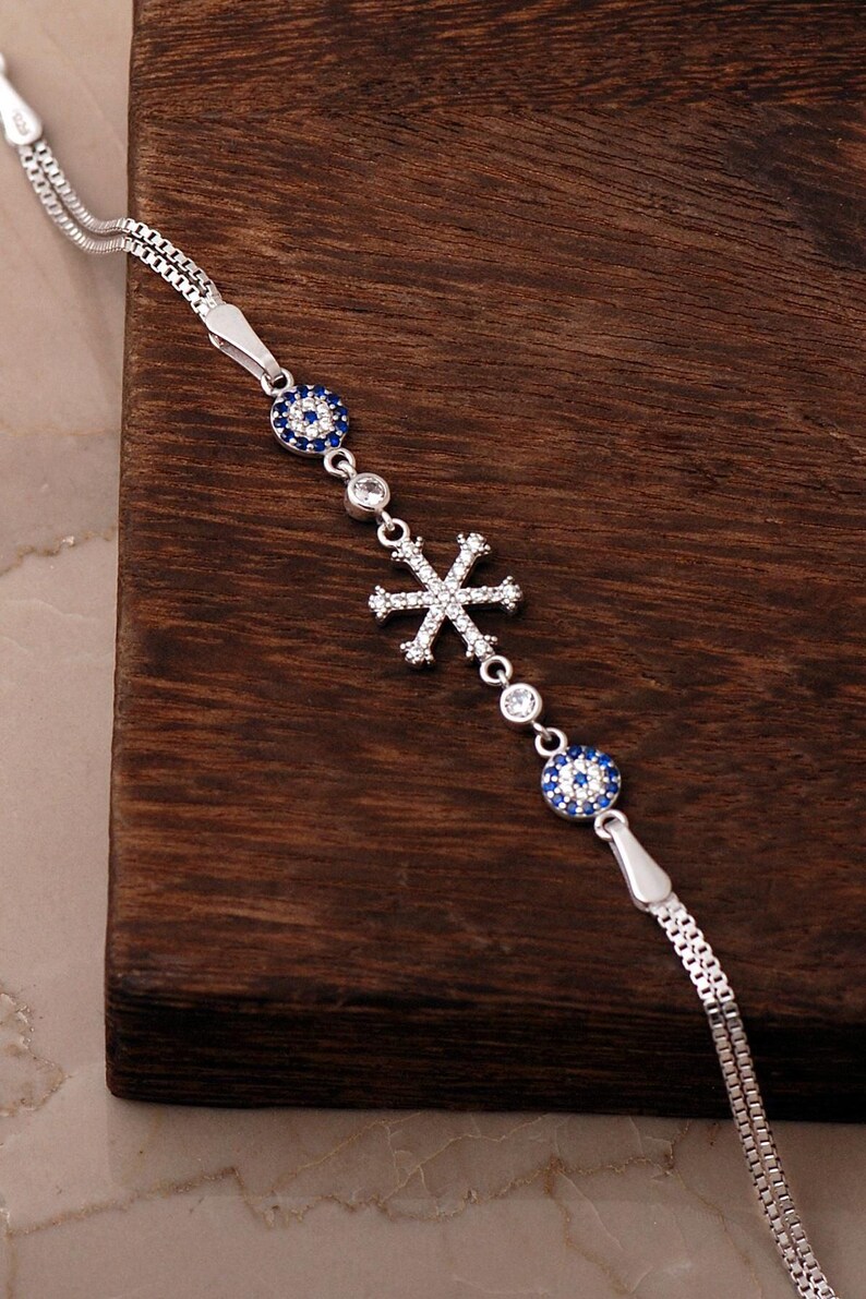 A delicate 925 sterling silver bracelet featuring a sparkling snowflake design combined with a blue sapphire and cubic zirconia evil eye charm on an adjustable box chain.