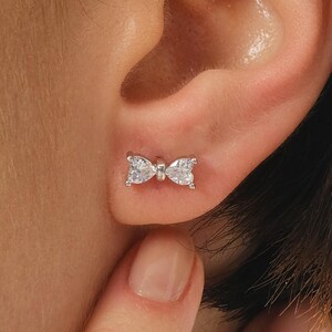 Mini bow‑shaped earrings made of rhodium‑plated sterling silver with a single white zircon stone on each side, measuring 4 mm tall by 1 cm wide.