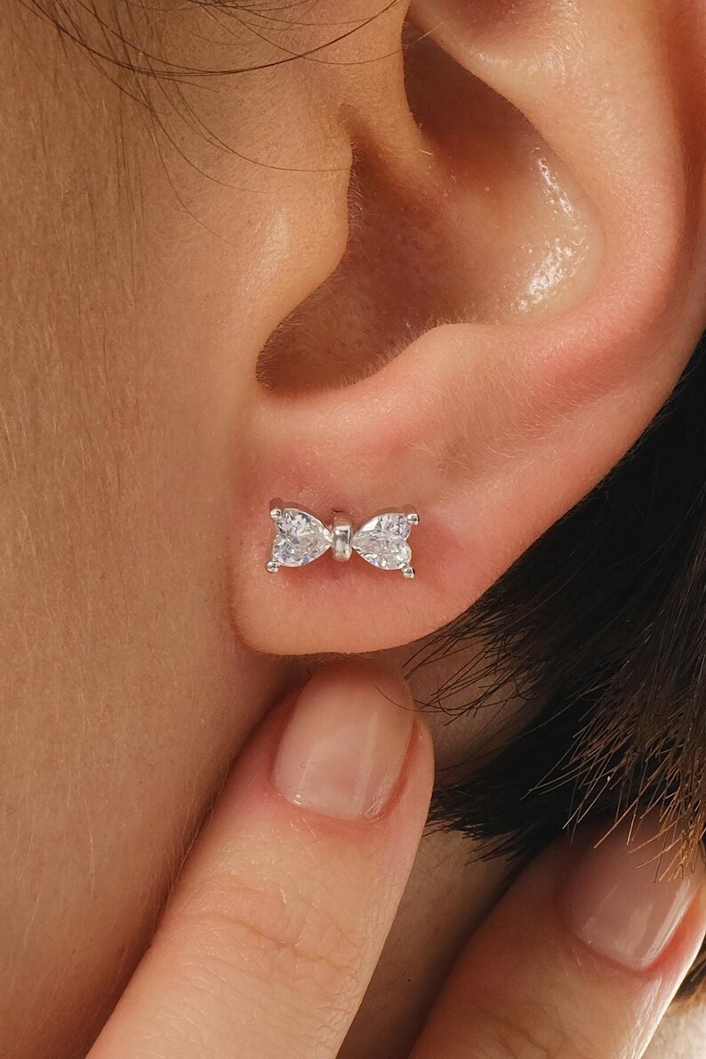 Mini bow‑shaped earrings made of rhodium‑plated sterling silver with a single white zircon stone on each side, measuring 4 mm tall by 1 cm wide.