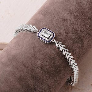 A detailed close-up of a 925 sterling silver herringbone chain bracelet featuring a dark blue enamel center piece adorned with a shimmering baguette-cut zircon stone.