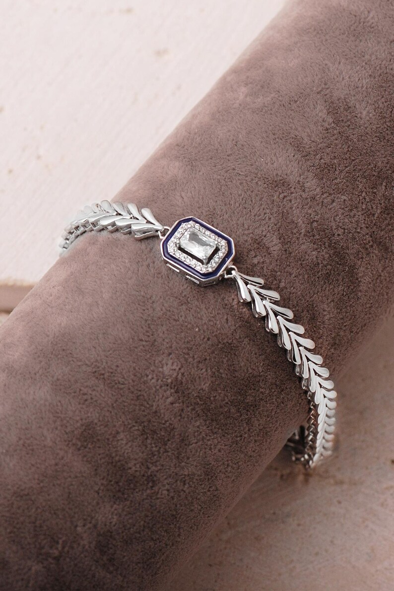 A detailed close-up of a 925 sterling silver herringbone chain bracelet featuring a dark blue enamel center piece adorned with a shimmering baguette-cut zircon stone.