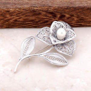 May include: A silver filigree flower brooch with a white pearl in the center. The flower has a delicate, intricate design and is accented by two leaves.