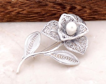 May include: A silver filigree flower brooch with a white pearl in the centre. The flower has a delicate, intricate design and is accented by two leaves.