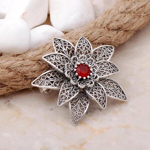 May include: A silver filigree flower brooch with a red gemstone in the center.