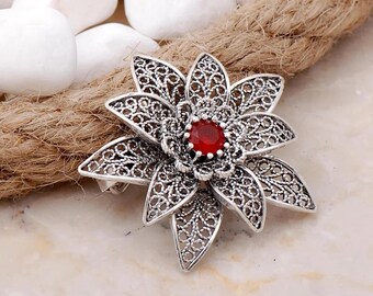 May include: A silver filigree flower brooch with a red gemstone in the centre.