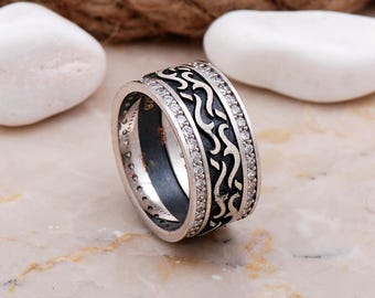 May include: A wide silver ring featuring a central band with a dark oxidized finish and a raised, flowing wave-like pattern. The central section is flanked by two thin rows of small, clear, round-cut stones set into the polished silver edges.