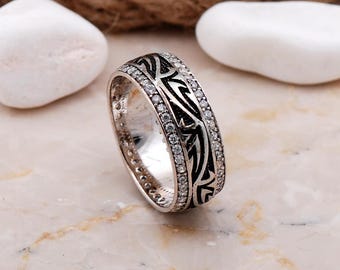 May include: A silver-toned ring featuring a central band with black engraved patterns, flanked by two rows of small, clear, round-cut stones. The metal has a polished finish, and the design includes intricate, dark, linear detailing.