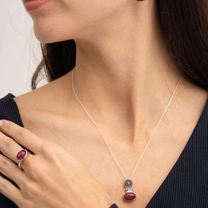 May include: A jewelry set consisting of a silver necklace with a pendant, matching drop earrings, and a ring. Each piece features a deep magenta oval gemstone set in silver with a circular, textured silver accent above the stone.