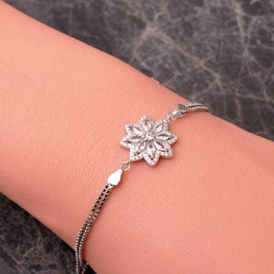 May include: A silver-toned bracelet featuring a central flower-shaped charm adorned with clear, marquise-cut stones. The bracelet is constructed with a delicate double-chain design, connecting to the floral centerpiece with small metal links.