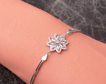 May include: A silver-toned bracelet featuring a central flower-shaped charm adorned with clear, marquise-cut stones. The bracelet is constructed with a delicate double-chain design, connecting to the floral centerpiece with small metal links.