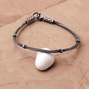May include: A silver bracelet featuring a woven chain design with oxidized detailing, small decorative knots along the band, and a lobster claw clasp. The bracelet is draped over a smooth, solid white stone with a rounded, irregular shape.