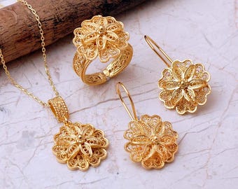 May include: A gold jewelry set consisting of a necklace with a flower-shaped pendant, a matching flower-shaped ring, and two flower-shaped drop earrings. Each piece features intricate filigree wirework with a delicate, open-work floral design.