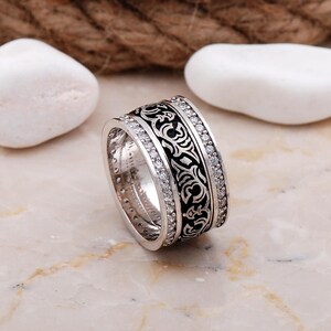 May include: A wide silver band ring featuring a central black inlay with an intricate engraved scrollwork pattern. The ring is bordered on both sides by two rows of small, clear, round-cut stones set into the polished silver metal surface.