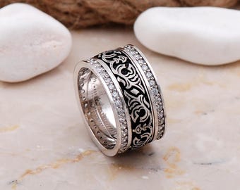 May include: A wide silver band ring featuring a central black inlay with an intricate engraved scrollwork pattern. The ring is bordered on both sides by two rows of small, clear, round-cut stones set into the polished silver metal surface.