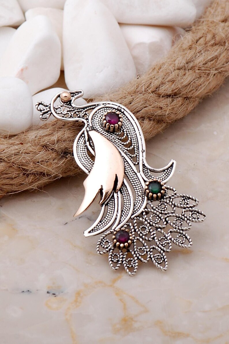 A handmade 925 sterling silver filigree brooch shaped like a peacock, featuring gold-toned accents and set with root ruby and emerald stones.