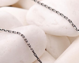 May include: A delicate, dark-toned metal chain necklace featuring small, uniform rectangular links. The chain is draped in a gentle curve, showcasing its oxidised finish and fine, lightweight construction throughout the entire length.