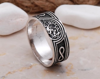 May include: A silver-toned metal ring featuring a wide band with intricate black engraved detailing. The surface displays a repeating pattern of infinity symbols and textured circular motifs, creating a high-contrast appearance.
