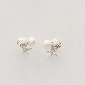 May include: A pair of silver-toned stud earrings featuring a small starfish and a ridged seashell design, accented with a single round white pearl. The earrings are arranged symmetrically, showcasing the detailed metallic texture of the sea-themed elements.