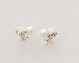 May include: A pair of silver-toned stud earrings featuring a small starfish and a ridged seashell design, accented with a single round white pearl. The earrings are arranged symmetrically, showcasing the detailed metallic texture of the sea-themed elements.