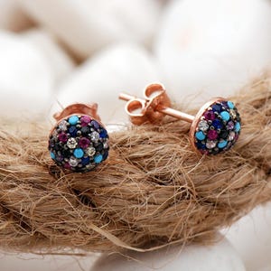 May include: A pair of rose gold stud earrings featuring a circular face encrusted with small, multicolored stones including turquoise, dark blue, magenta, and clear white gems. Each earring has a standard post back closure for secure attachment.
